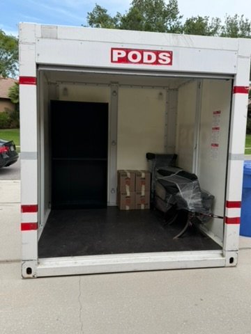 An open PODS moving and storage container parked on a residential street, partially loaded with wrapped furniture, boxes, and a large black appliance.