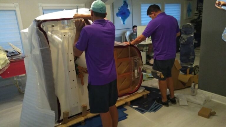 Two movers padding a baby grand piano on its side on a piano board in South Tampa, FL